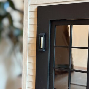 Op de afbeelding: Een miniatuur zwarte voordeur met een meerruits raam en een bijpassende wandlamp. De deur staat tegen een achtergrond van lichtgekleurde gevelbekleding, wat een realistisch architectonisch detail cre&euml;ert.