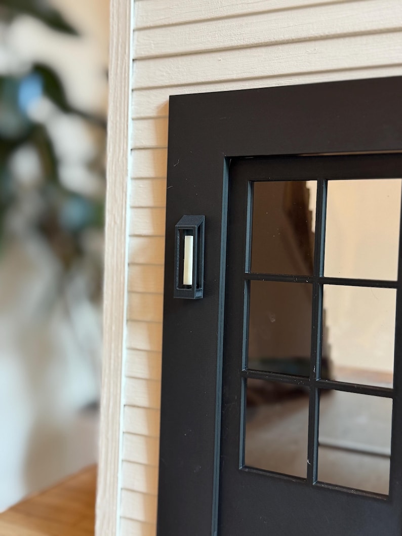 Op de afbeelding: Een miniatuur zwarte voordeur met een meerruits raam en een bijpassende wandlamp. De deur staat tegen een achtergrond van lichtgekleurde gevelbekleding, wat een realistisch architectonisch detail cre&euml;ert.