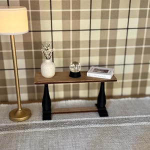 May include: A miniature living room scene featuring a small wooden table with black legs, a white vase with sprigs, a crystal ball, and a book titled "The Art of Home." A gold floor lamp stands to the left. The background is a plaid pattern.