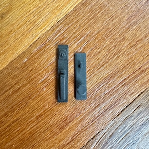 May include: Two small, rectangular, matte black plastic components resembling miniature door hardware. The left piece features a circular top detail and a vertical handle, while the right piece displays a small central protrusion and a circular base.