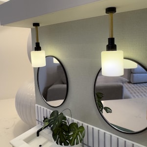 Könnte beinhalten: Ein modernes Badezimmer-Waschbecken mit zwei Waschbecken, weißen Arbeitsplatten und einer Pflanze. Zwei runde Spiegel hängen über den Waschbecken, jeweils mit einer mattierten Glaspendelleuchte darüber. Die Wände sind mit weißen und grauen Fliesen gefliest.