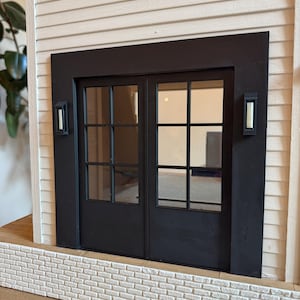 Op de afbeelding: Een miniatuur zwarte dubbele deur met glazen ruiten, geplaatst in een zwart frame. De deur wordt geflankeerd door twee rechthoekige wandkandelaars. De structuur heeft witte gevelbekleding en een bakstenen gevel, waardoor een gedetailleerd architectonisch model ontstaat.