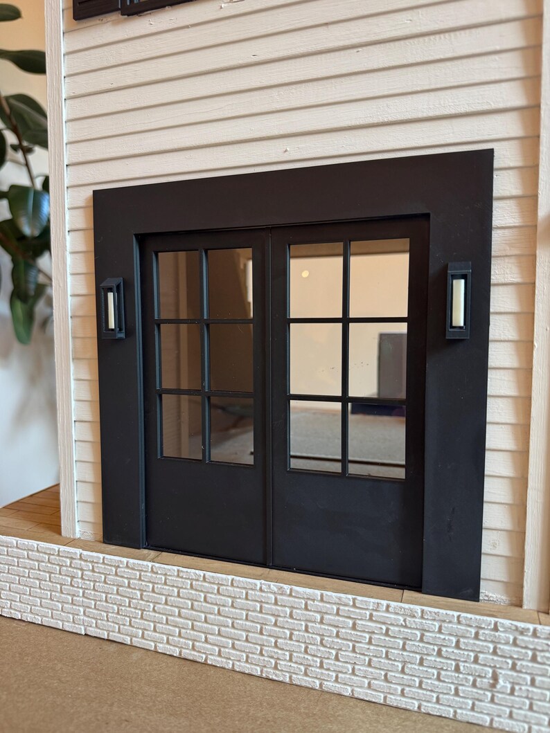 Op de afbeelding: Een miniatuur zwarte dubbele deur met glazen ruiten, geplaatst in een zwart frame. De deur wordt geflankeerd door twee rechthoekige wandkandelaars. De structuur heeft witte gevelbekleding en een bakstenen gevel, waardoor een gedetailleerd architectonisch model ontstaat.