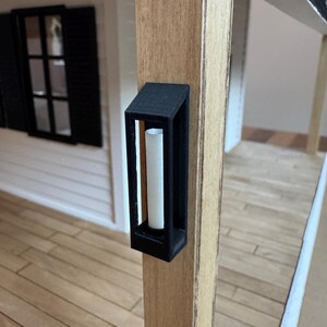 Op de afbeelding: Een zwarte miniatuur buitenwandlamp met een witte kaars aan de binnenkant is gemonteerd op een houten paal. De lamp heeft een rechthoekig frame en maakt deel uit van een poppenhuisveranda. De veranda heeft een witte buitenkant en zwarte luiken.
