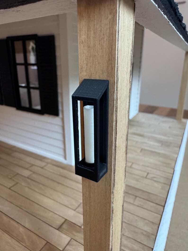 Op de afbeelding: Een zwarte miniatuur buitenwandlamp met een witte kaars aan de binnenkant is gemonteerd op een houten paal. De lamp heeft een rechthoekig frame en maakt deel uit van een poppenhuisveranda. De veranda heeft een witte buitenkant en zwarte luiken.