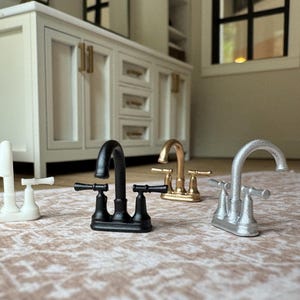 May include: Four miniature bathroom sink faucets in white, matte black, gold, and silver finishes. Each faucet features a curved spout and two horizontal handles on a rectangular base, designed as small-scale replicas for dollhouse decor.