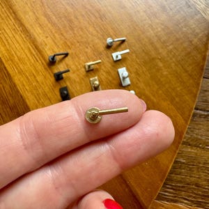 May include: A miniature gold-toned metal door handle with a circular base and a straight lever. The handle features a textured, beaded edge around the circular plate. Several other miniature handles in black, gold, and silver finishes are visible.