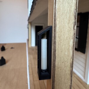 Op de afbeelding: Een miniatuur zwarte lantaarn met een witte kaars erin is gemonteerd op een houten paal. De lantaarn heeft een rechthoekig frame en maakt deel uit van een poppenhuis of modelhuis.