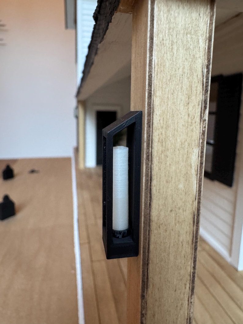 Op de afbeelding: Een miniatuur zwarte lantaarn met een witte kaars erin is gemonteerd op een houten paal. De lantaarn heeft een rechthoekig frame en maakt deel uit van een poppenhuis of modelhuis.