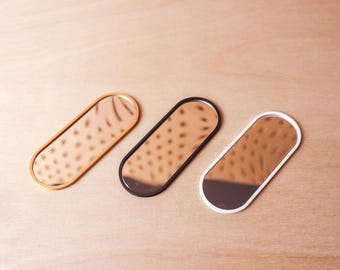 May include: Three oval-shaped mirrors with different colored frames: gold, black, and white. The mirrors have a reflective surface with a dotted pattern. The mirrors are arranged on a light brown wooden surface.