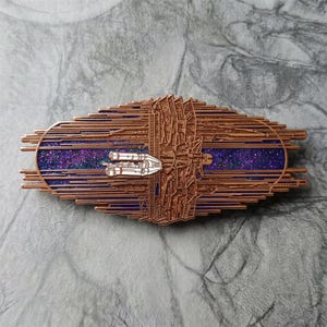 May include: A copper-coloured metal pin with a space theme. The pin features a detailed spaceship design, a starry purple background, and a geometric structure. The pin is an oval shape with a horizontal orientation.