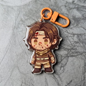 Puede incluir: Un llavero de dibujos animados con un personaje masculino de pelo castaño, una cinta en la cabeza y barba. El personaje lleva un atuendo marrón y tiene sangre en la cara. El llavero tiene un cierre naranja.