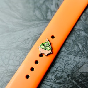 Può includere: Un cinturino arancione con una spilla decorativa. La spilla presenta un design a foglia verde con uno sfondo rosa chiaro. Il cinturino ha diversi fori per la regolazione.