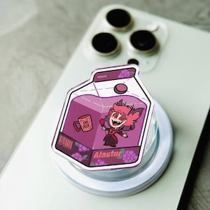 Puede incluir: Un soporte de teléfono acrílico transparente con un diseño de personaje de dibujos animados dentro de una caja de jugo púrpura. La caja de jugo tiene una parte superior blanca y un fondo púrpura con el texto "Alastor".