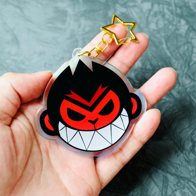 May include: A clear acrylic keychain featuring a cartoon monkey face with a red face, black hair, and white teeth. The keychain has a gold star-shaped clasp and a gold chain. The monkey has an angry expression.