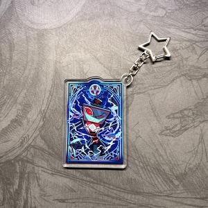 May include: A rectangular acrylic keychain featuring a cartoon character with a red and blue color scheme. The character is set against a blue background with white lightning-like patterns. A silver star-shaped charm is attached.