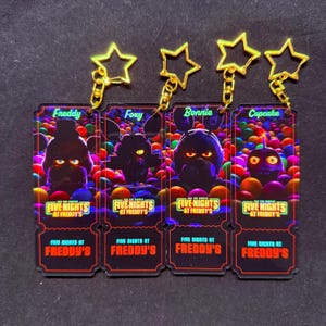 May include: Four colorful keychains featuring characters from the video game Five Nights at Freddy's. Each keychain has a gold star charm and a black and red ticket-shaped charm with a character's name and image. The characters are Freddy, Foxy, Bonnie, and Cupcake.