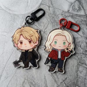 May include: Two acrylic keyrings with cartoon character illustrations. One has a black clasp and shows a character with blonde hair, a black jacket, and an axe. The other has a red clasp and depicts a character with blonde hair, a red jacket, and a gun.
