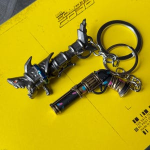 May include: Two keychains, one shaped like a futuristic gun and the other shaped like a pistol. The gun keychain is silver with blue accents and the pistol keychain is black with pink and blue accents.