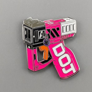 Puede incluir: Un pin de esmalte rosa y negro con forma de pistola futurista. El pin presenta la palabra "DOT" en letras blancas, junto con otros diseños geométricos y la letra "Y". Un pequeño dije morado cuelga del pin.