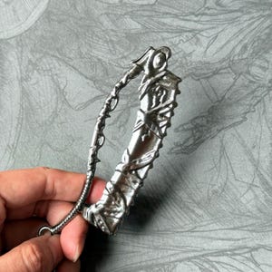 Op de afbeelding: Een zilverkleurige metalen sleutelhanger met een gedetailleerd ontwerp van een gebogen mes met een gekartelde rand. De sleutelhanger heeft een ring aan de bovenkant om aan sleutels te bevestigen.