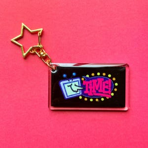 May include: A rectangular keychain with a gold star-shaped clasp. The keychain features a black background with a cartoon television and the word "TIME!" in pink and yellow. The TV has a smiling face.