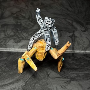 May include: A detailed sculpture of a figure atop a multi-legged creature. The figure is silver, with an open, geometric design. The creature is primarily tan with blue accents. The artwork is set against a dark, patterned background.