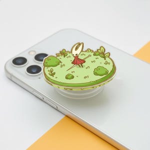 Puede incluir: Un soporte para teléfono verde y blanco con un personaje de dibujos animados con un vestido rojo, de pie sobre una zona de hierba verde con arbustos. El accesorio está fijado a un smartphone plateado.