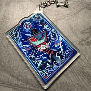 May include: A rectangular acrylic keychain featuring a stylized character design. The design is primarily blue, red, and white, with a detailed illustration of a character with a television-like head. The keychain has a silver chain and clasp.