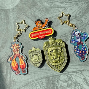 May include: Five gold-coloured metal keychains featuring characters from the video game Five Nights at Freddy's. The keychains are on a grey surface. The keychains feature Freddy Fazbear, a security guard, a clown, and two other characters.