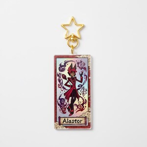 May include: A rectangular keychain featuring a character illustration with the name "Alastor" at the bottom. The character is depicted in a red and black outfit, surrounded by purple and black swirls. The keychain has a gold star-shaped clasp.