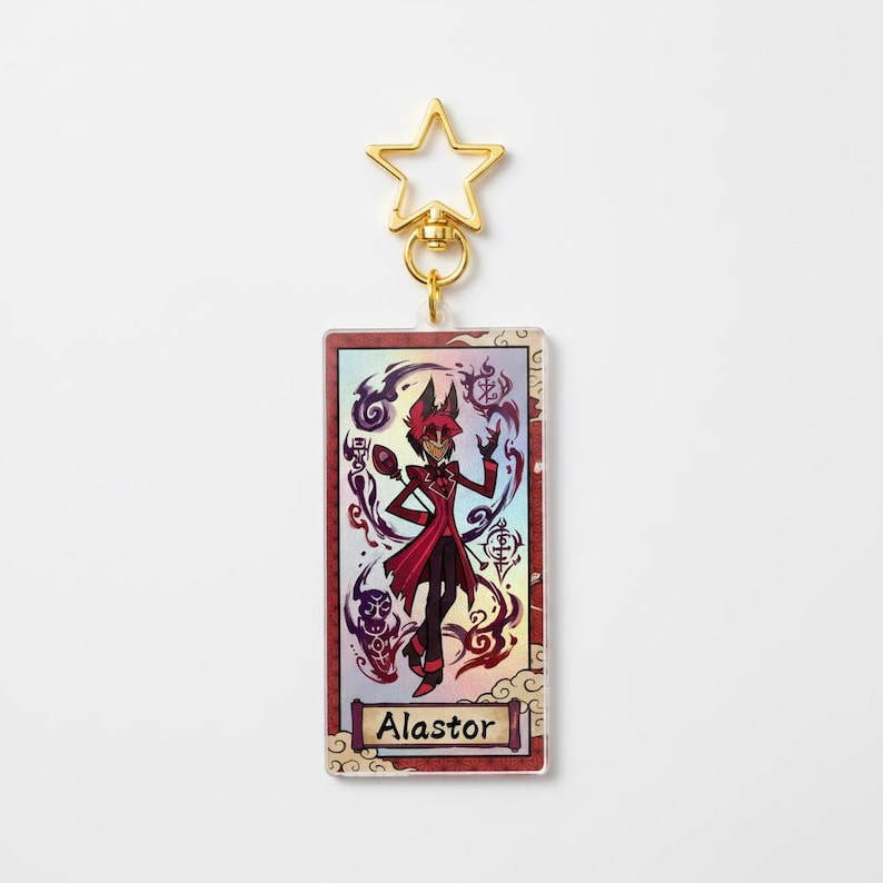 May include: A rectangular keychain featuring a character illustration with the name "Alastor" at the bottom. The character is depicted in a red and black outfit, surrounded by purple and black swirls. The keychain has a gold star-shaped clasp.