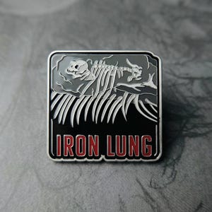 May include: Square enamel pin with a silver border. The pin features a grey skeletal design over a black background. The text "IRON LUNG" is in red letters at the bottom of the pin.