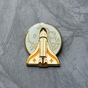 K&ouml;nnte beinhalten: Eine goldfarbene Emaille-Anstecknadel mit einem wei&szlig;en Raumschiff mit orangefarbenen Akzenten, das vor einem wei&szlig;en Mond mit grauen Kratern fliegt.