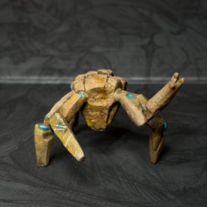 May include: A handcrafted, stylized sculpture of a creature with a segmented body and legs. The sculpture is primarily brown with gold accents and small blue details. The creature's pose suggests movement, with one arm raised.