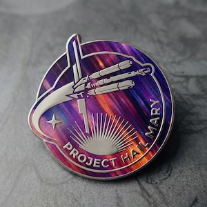Pode incluir: Um alfinete de metal redondo prateado com um fundo colorido e rodopiante de roxo, laranja e azul. O alfinete apresenta um design estilizado de nave espacial, uma estrela e o texto "PROJECT HALL MARY".