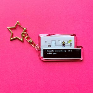 May include: A clear acrylic keychain with a gold star-shaped clasp. The keychain features a pixelated scene with a character and the text "Despite everything, it's still you."