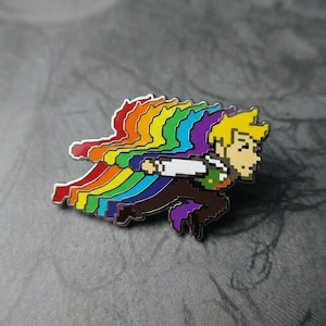 Op de afbeelding: Een metalen emaille speld met een pixel art personage in een rennende houding, met een levendig regenboogeffect erachter. Het personage heeft blond haar, een wit overhemd, een groen vest en een bruine broek, met regenboogkleuren in lagen.