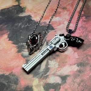 Puede incluir: Dos collares de plata con colgantes. Un colgante es una pistola con un cañón plateado y un mango negro. El otro colgante es un diseño ornamentado plateado y rojo con una piedra preciosa roja.