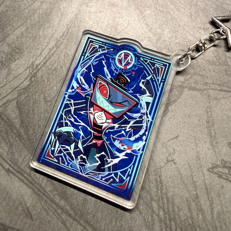 May include: A rectangular acrylic keychain featuring a cartoon character with a screen-shaped head, red eyes, and a sharp-toothed grin. The design is set against a blue background with white lightning-like patterns and a decorative border. A silver chain and clasp are attached.