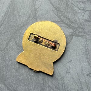K&ouml;nnte beinhalten: Die R&uuml;ckseite einer goldfarbenen Anstecknadel mit Sicherheitsverschluss. Die Nadel ist rund mit einem erh&ouml;hten Rand und einer glatten Oberfl&auml;che.