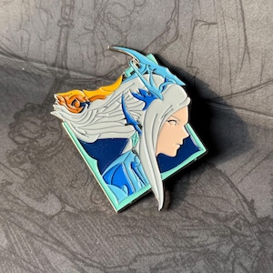 Puede incluir: Un pin de esmalte azul y blanco con un personaje de pelo largo blanco y un diseño de dragón azul y blanco. El personaje lleva un atuendo azul y blanco y tiene un fondo azul.