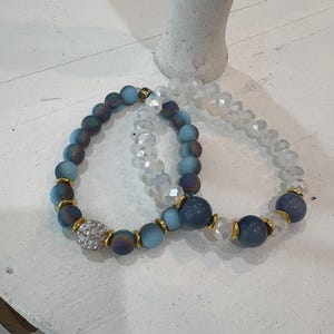 以下が含まれることがあります： 2つのブレスレット。1つは虹色の青いビーズとキラキラ光る銀色のチャーム、もう1つは透明なクリスタルビーズと青いビーズのチャームが付いている。