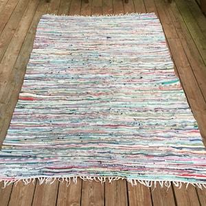 Pu&ograve; includere: Un tappeto rettangolare multicolore di stracci con strisce orizzontali di vari colori tra cui bianco, blu, rosso e verde. Il tappeto ha un bordo sfrangiato ed &egrave; esposto su un terrazzo di legno.