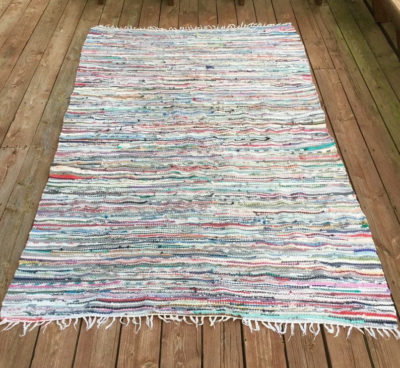 Pu&ograve; includere: Un tappeto rettangolare multicolore di stracci con strisce orizzontali di vari colori tra cui bianco, blu, rosso e verde. Il tappeto ha un bordo sfrangiato ed &egrave; esposto su un terrazzo di legno.