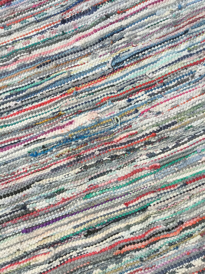 Pu&ograve; includere: Primo piano di un tappeto colorato e tessuto con un motivo a righe diagonali. Il tappeto presenta un mix di colori tra cui grigio, bianco, rosso, blu e verde. La trama sembra essere fatta di strisce di tessuto.