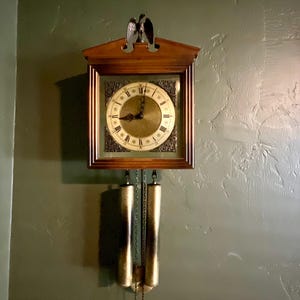 May include: A wooden wall clock featuring a square frame, a circular face with Roman numerals, and decorative corner accents. An eagle figure sits atop the clock, and two cylindrical brass-toned weights hang from chains below the main housing.