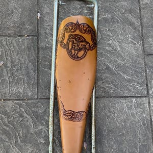 Könnte beinhalten: Ein brauner Ledersattel für ein Fahrrad mit einem dekorativen Design eines Cowboys, der ein Pferd reitet. Der Sattel ist an einem Metallrahmen befestigt.