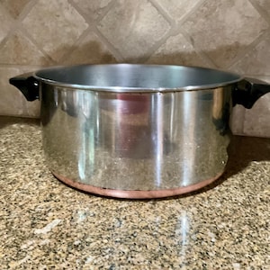 Puede incluir: Una olla redonda de acero inoxidable con un acabado plateado pulido y una base de color cobre visible. La olla cuenta con dos asas de plástico negro resistentes al calor, fijadas en los lados opuestos del borde para un agarre seguro.