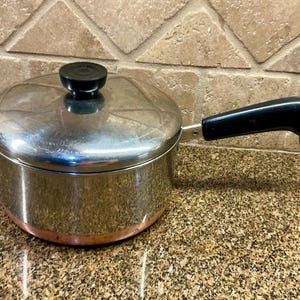 May include: A stainless steel saucepan with a black handle and lid. The pot has a copper-coloured bottom and a black knob on the lid. The handle has a metal loop at the end.